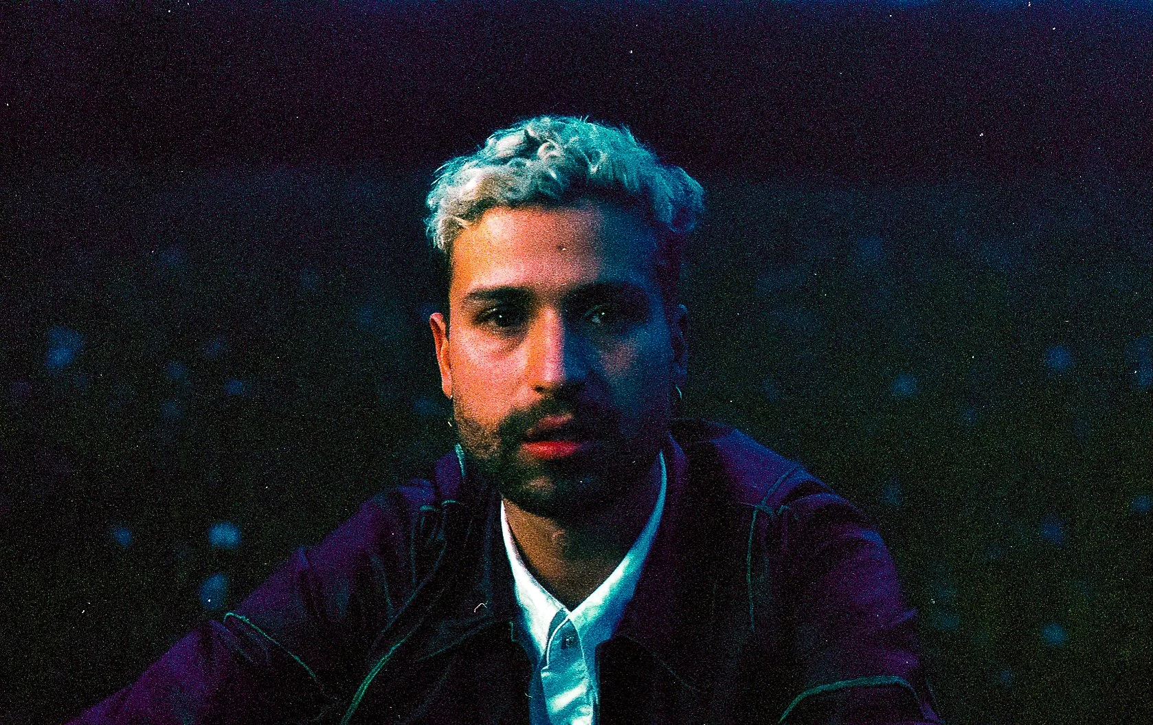 A young man with blonde,/or platinum hair, wearing a dark jacket and white shirt, looking directly at the camera with a serious expression, against a dark, starry background illuminated with colored lights.