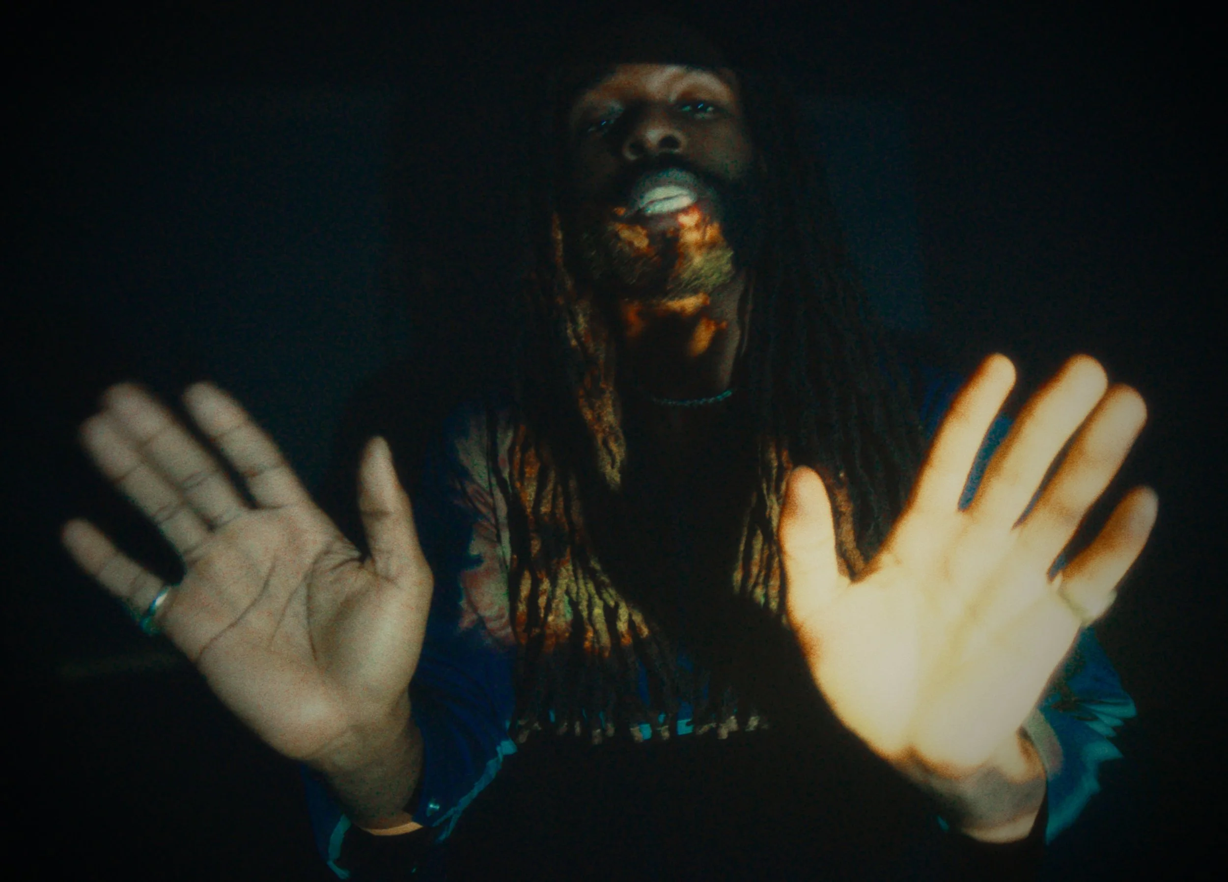 A person with long dreadlocks, wearing a dark top, holding their hands up towards the camera, with a dark background.