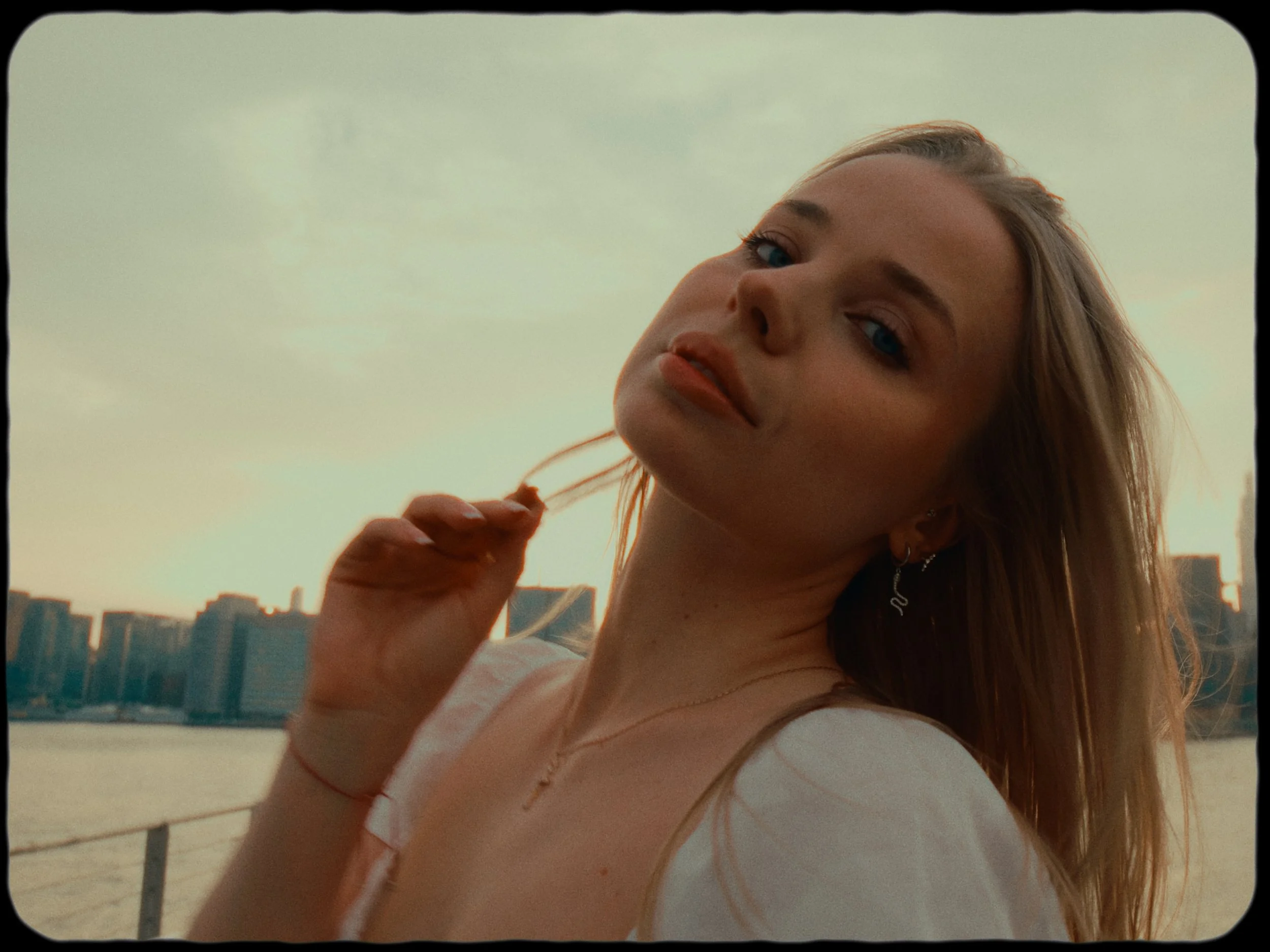 A young woman with blonde hair, blue eyes, and earrings, standing outdoors near a body of water with city skyline in the background during sunset.