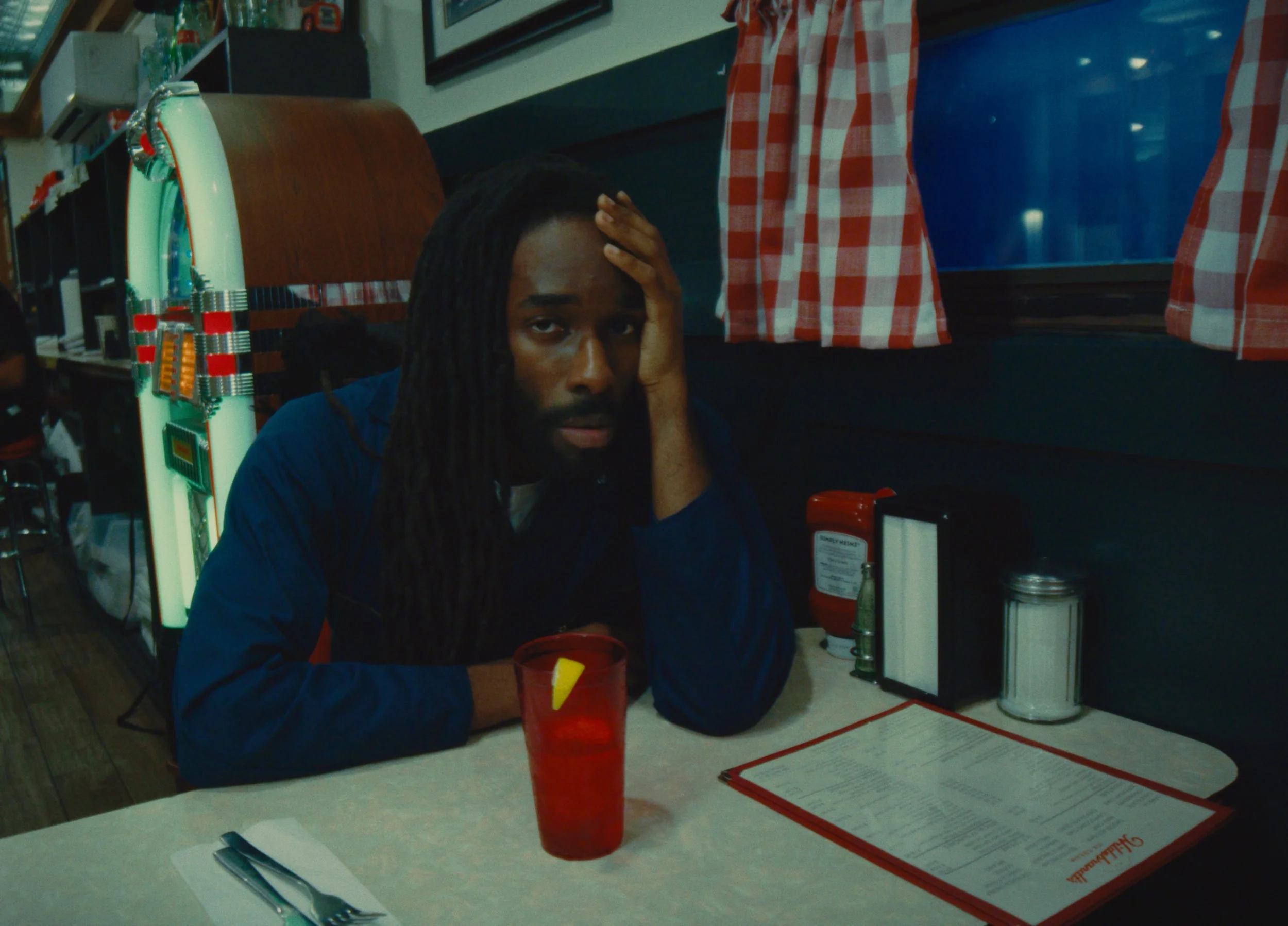 A man sitting alone at a restaurant table appears bored or upset, resting his head on his hand, wearing a dark jacket, with a red drink and a menu on the table.