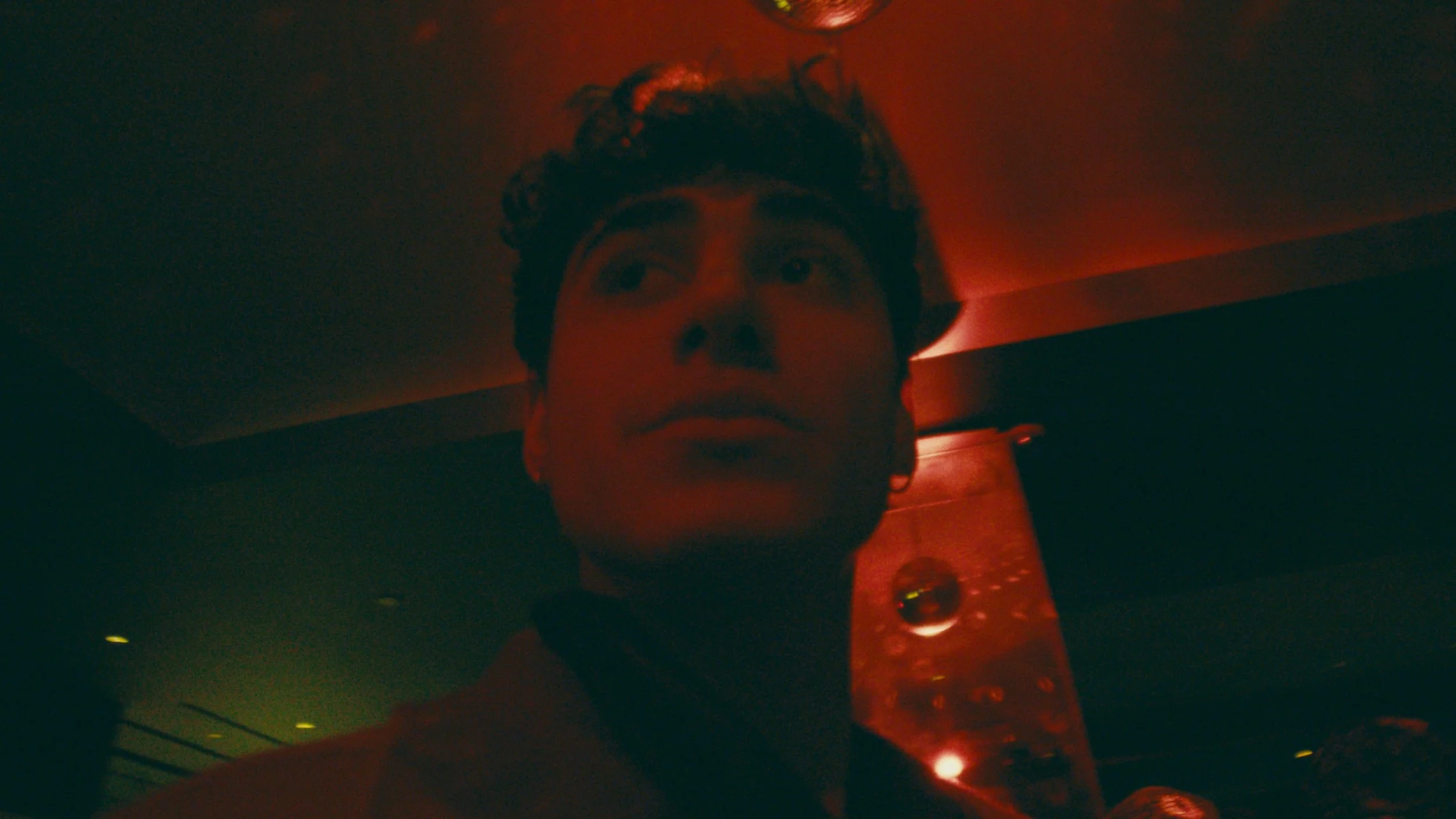 Close-up of a young man with curly hair, wearing an earring, in a dimly lit room with red lighting.