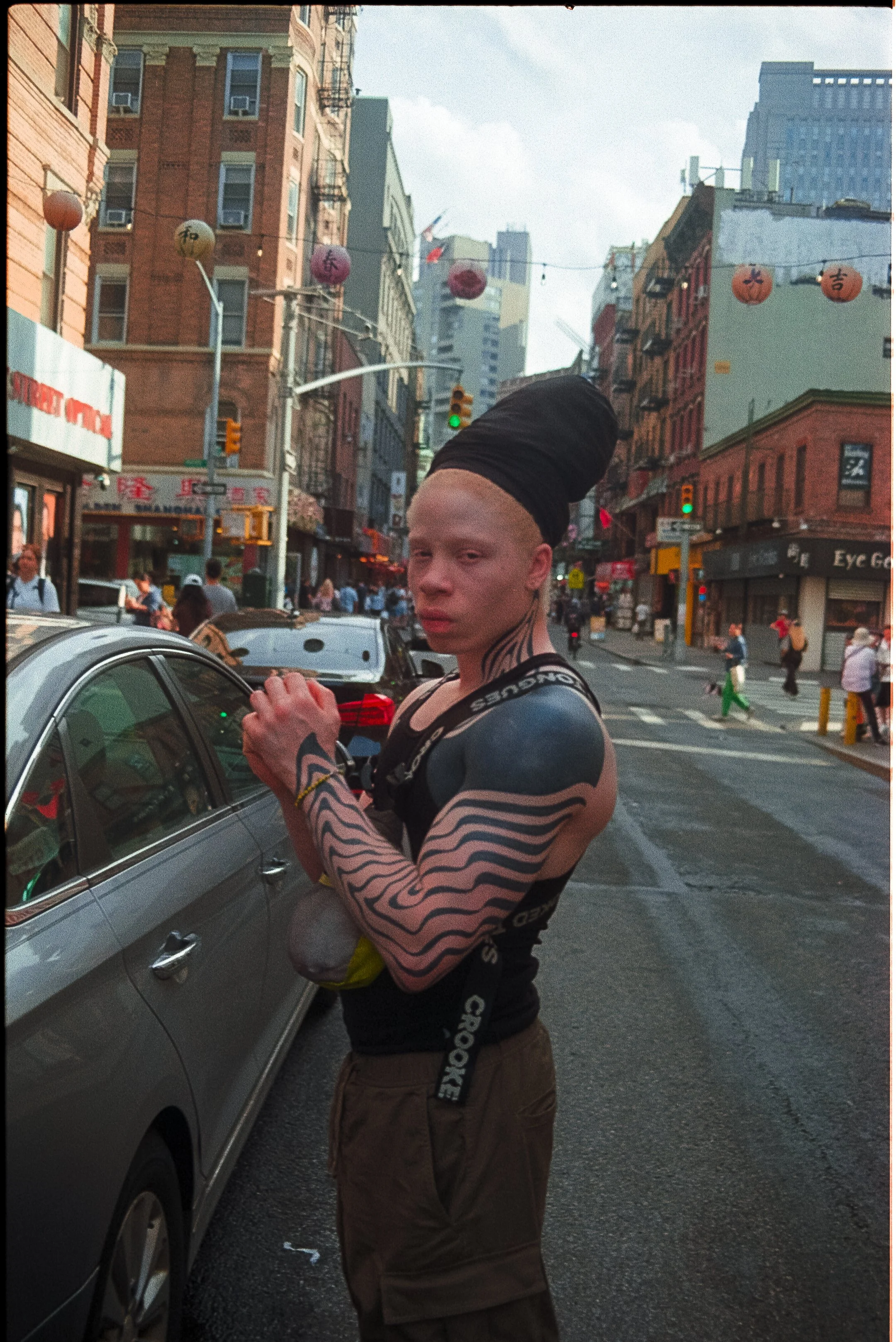 A person with tattoos on their arm and neck, wearing a black hood and a black tank top, standing on a city street holding a shopping bag. The background shows buildings, pedestrians, cars, and hanging lanterns.