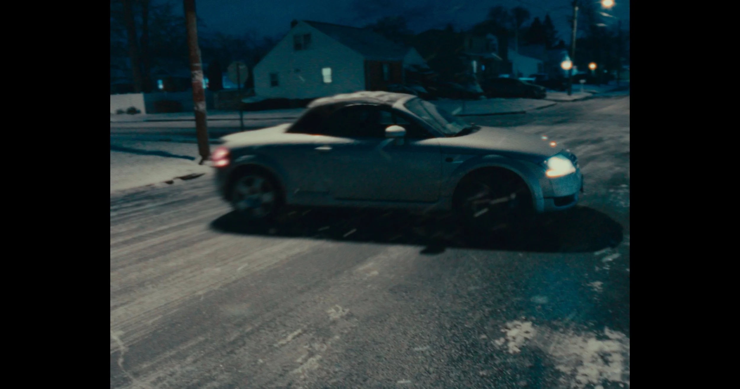 A silver convertible car parked on a snowy road at night with houses and streetlights in the background.