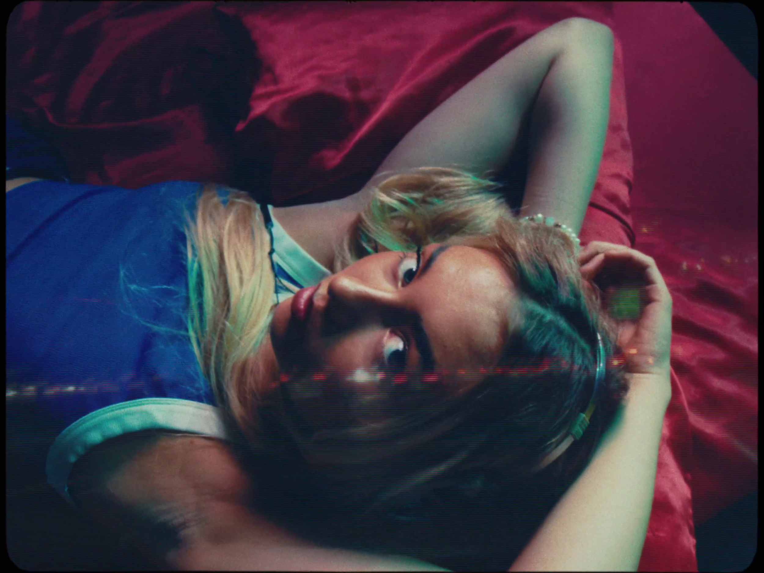 A woman with blonde hair and glasses lying on a bed with red sheets, looking at the camera.