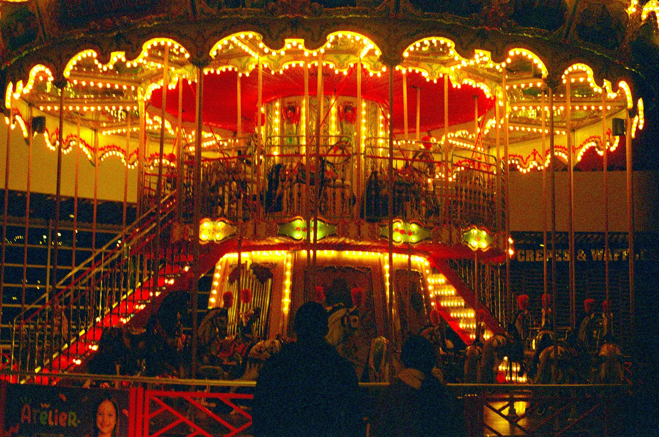 Nighttime photo of a brightly lit, double-decker carousel with children riding on the lower level. The carousel is decorated with numerous lights and painted panels.