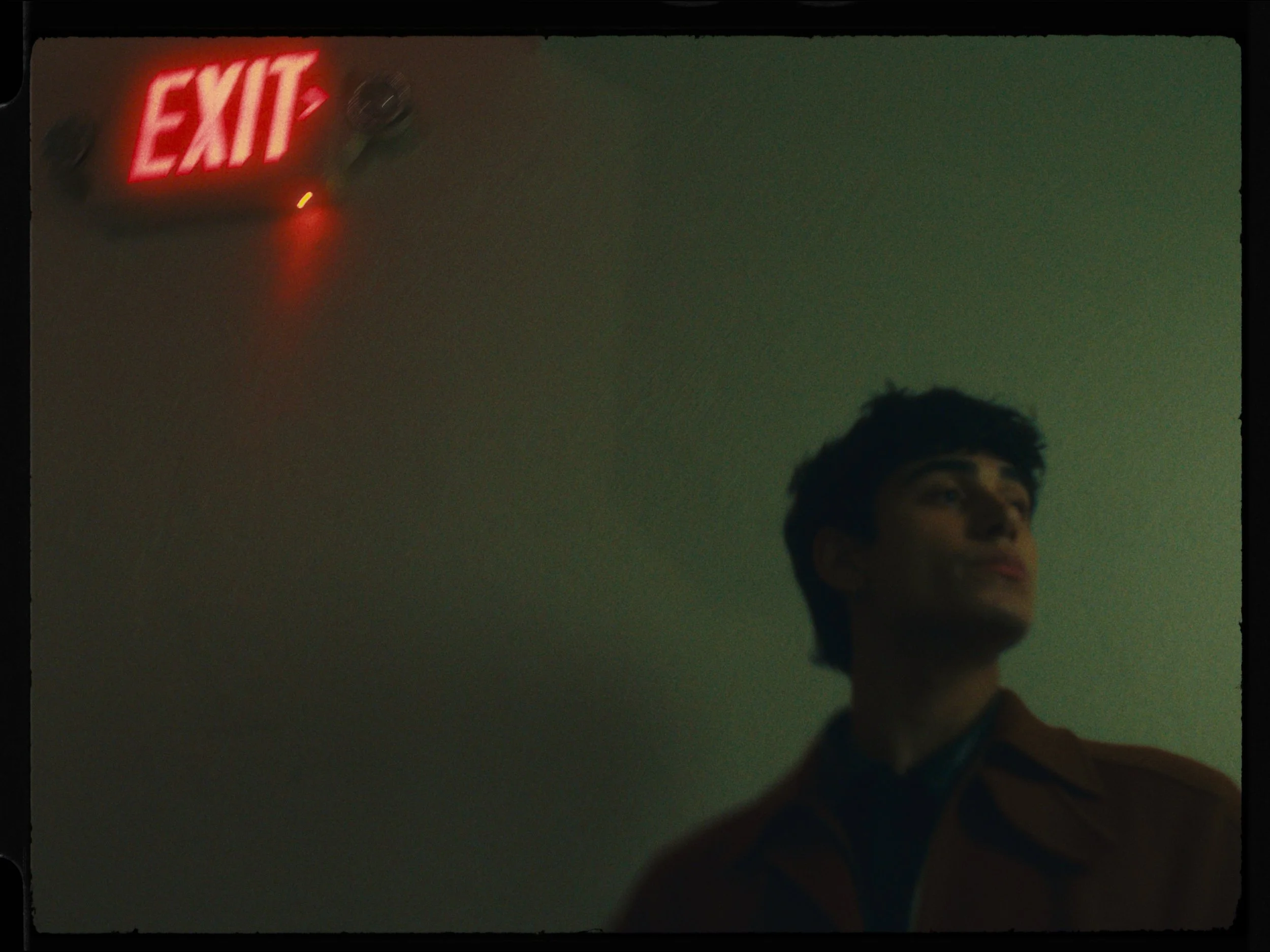Young man with dark hair in a brown jacket looking to the side in a dimly lit room with a red illuminated exit sign on the ceiling.