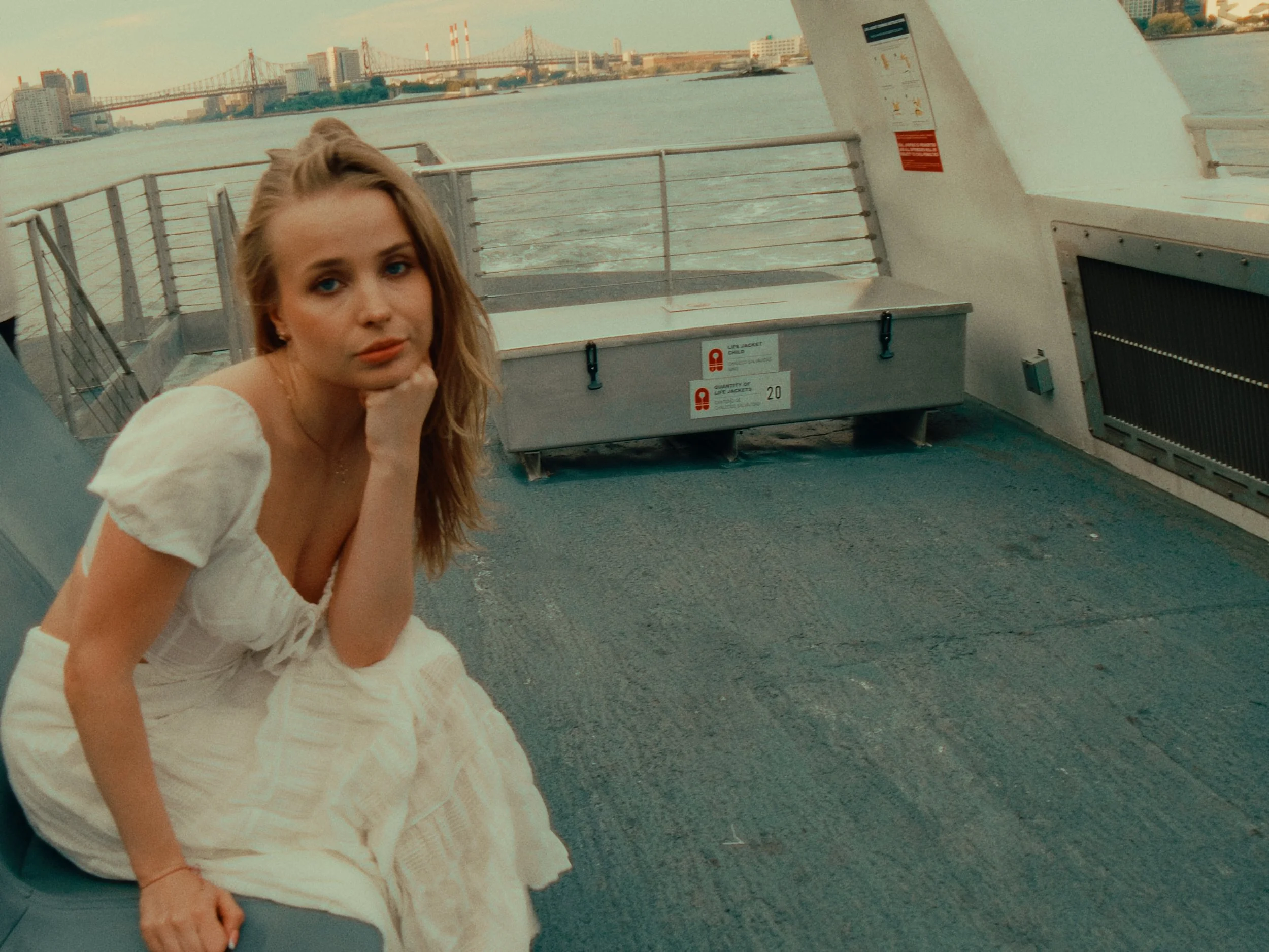 A young woman sitting on a boat deck, resting her chin on her hand and looking at the camera, with city buildings and a bridge in the background.