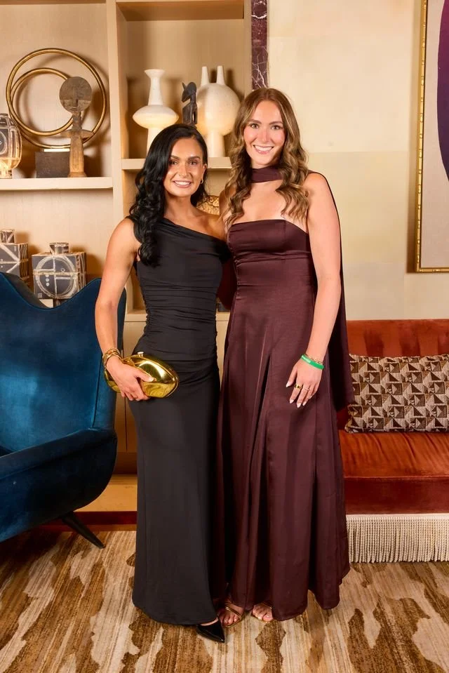 Two women dressed in elegant evening gowns standing indoors with decorative vases and artwork in the background.