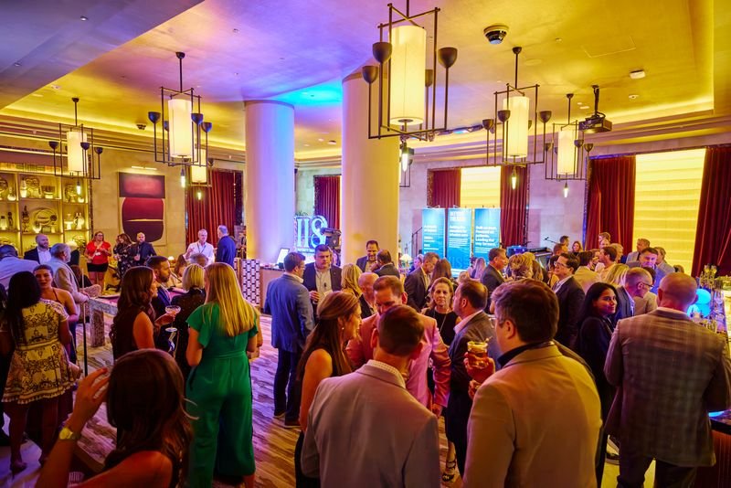 A crowded indoor party or social event with people mingling and talking. The room has modern lighting, tall white columns, red curtains, and warm decor.