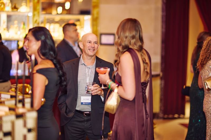 People socializing at an indoor event or party, holding drinks, with a bar in the background.