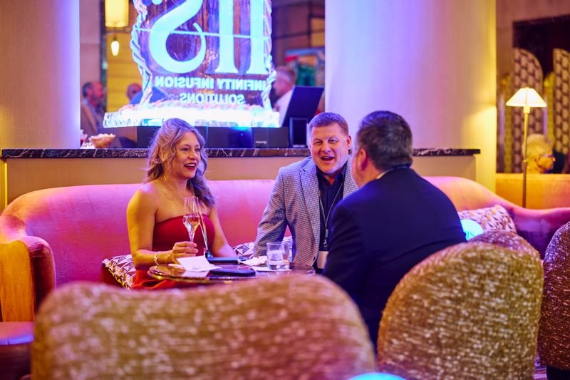 Three people sitting on a pink sofa and talking in a restaurant or lounge, with a neon sign in the background, and drinks on the table.
