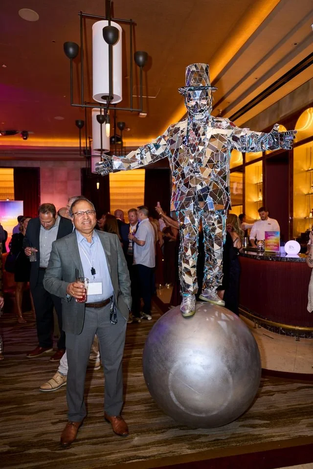 Man in a gray suit with glasses standing with a drink in a hotel lobby or conference room, next to a large sphere sculpture with a mirrored disco ball figure of a man in a suit and hat balancing on top, arms outstretched, with people in the backgroun