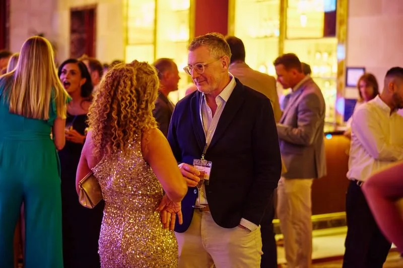 People socializing at a party or event in a warmly lit venue, with a focus on a man and woman talking in the foreground.