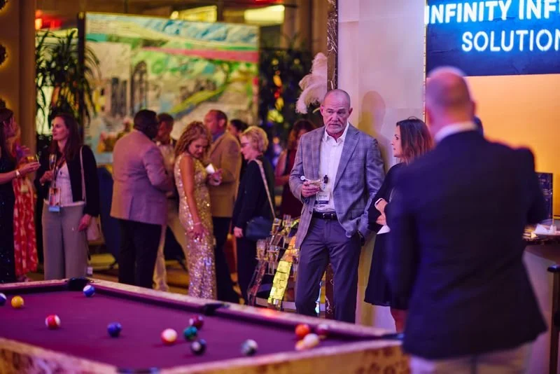 A group of people at a social event or party, with a pool table in the foreground and a digital screen on the wall displaying 'INFINITY INFINITY SOLUTIONS' in the background.