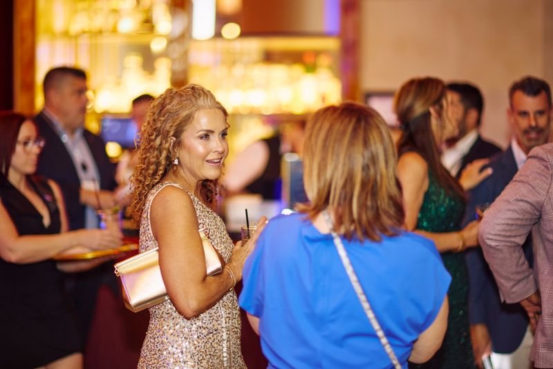 Group of people socializing at a bar or lounge, with a woman in a sparkly dress holding a drink in the foreground.