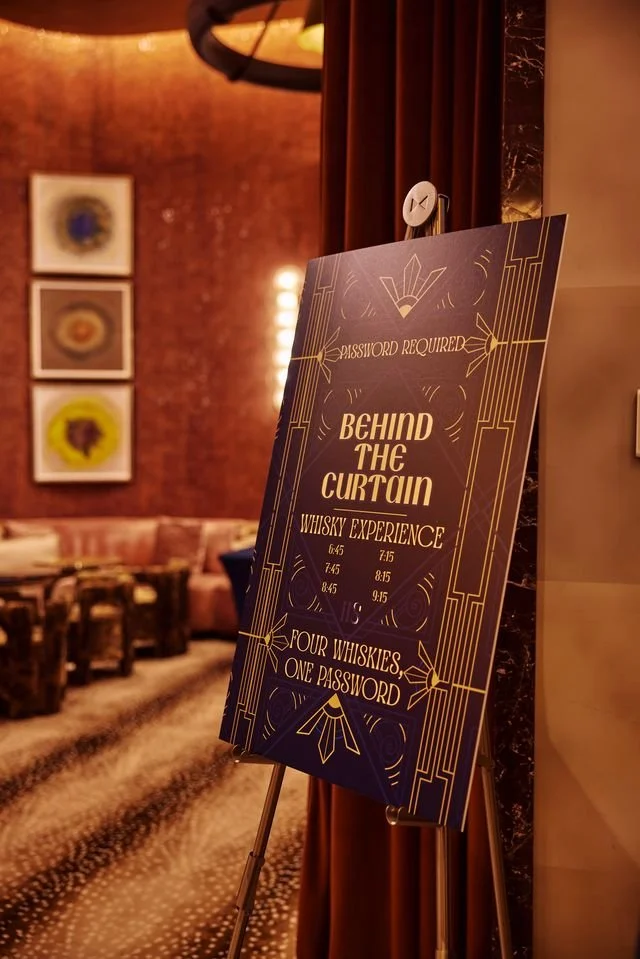 A signboard in a dimly lit bar or lounge reads 'Behind the Curtain Whisky Experience' and 'Four Whiskies, One Password'. The sign mentions that a password is required and lists several times for the experience, with an elegant art deco design. The ba