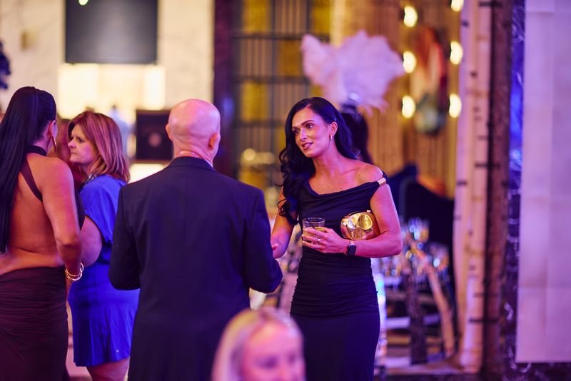 A group of people at a social event, with three women and one man engaged in conversation. The woman on the right is holding a drink and a clutch purse, wearing a black off-shoulder dress. The background shows warm lighting and decorative feathers.