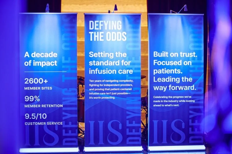 Three blue banners at an event, displaying texts about a decade of impact, setting standards for infusion care, and trust in a healthcare industry, with a stage and chairs in the background.