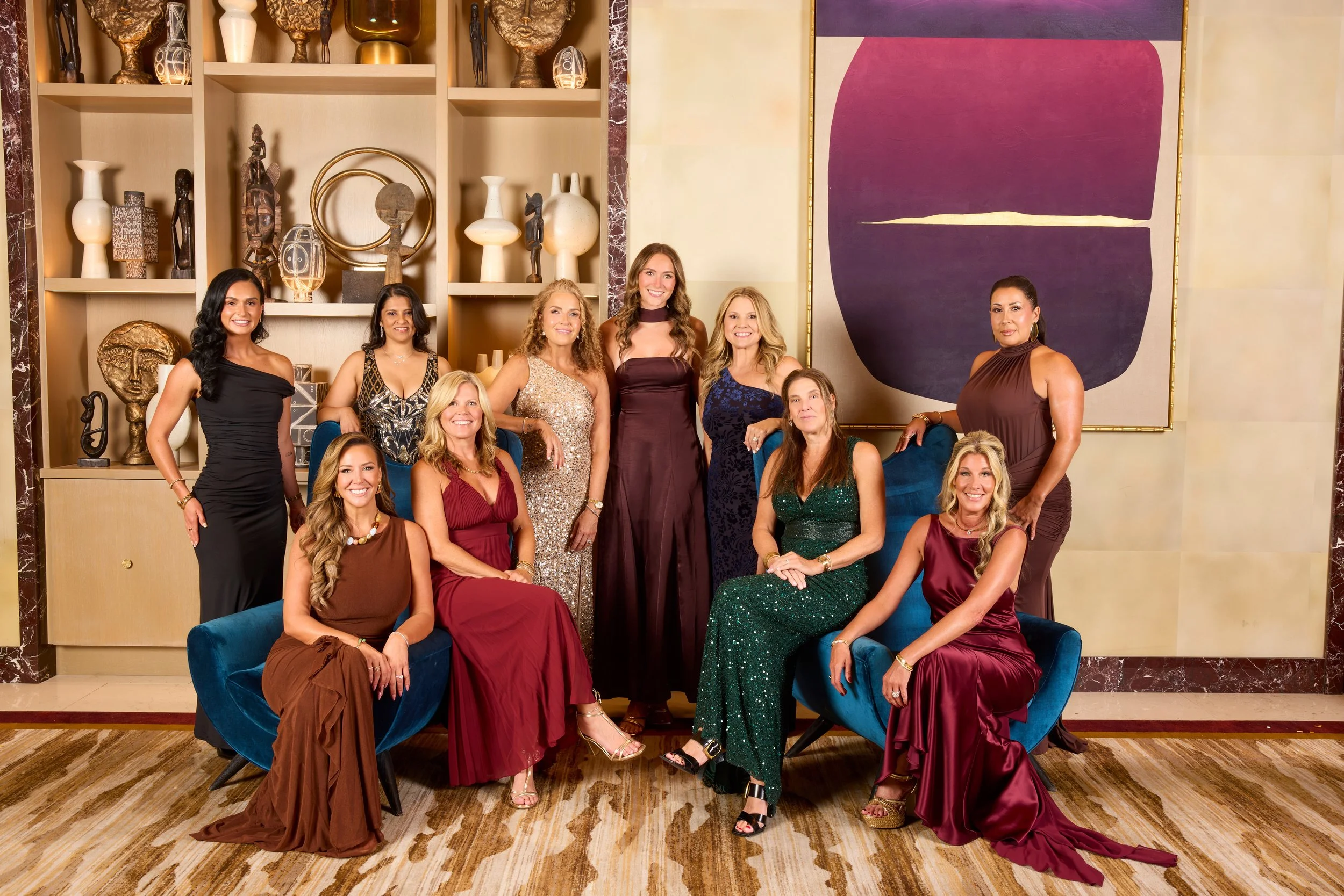 A group of women dressed in formal evening gowns posing in a luxurious hotel lobby with decorative sculptures, vases, and modern artwork on the wall behind them.
