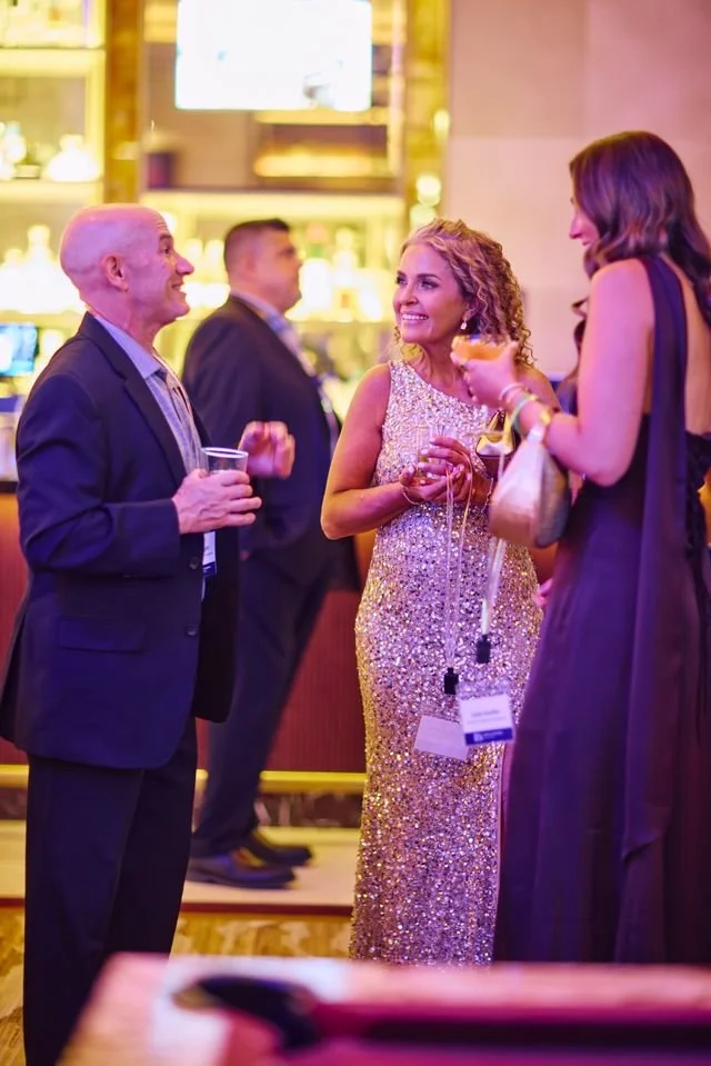 People socializing at an indoor event, with one man holding a drink and engaged in conversation with two women, one in a sparkly dress and the other in a dark dress, in front of a bar with warm lighting.