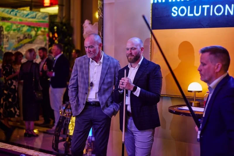 Three men in business suits stand near a wall with a large blue screen, one holding a microphone, at a business event or conference with other attendees in the background.