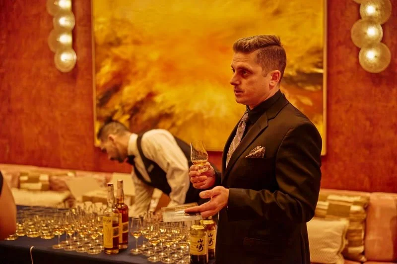 Man in a black suit holding a glass of whiskey at a tasting event with bottles and glasses on a table behind him, and a server in the background in a restaurant or lounge with warm lighting and abstract wall art.