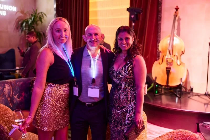 Three people standing together at a social event, smiling, with a cello on a stand in the background.