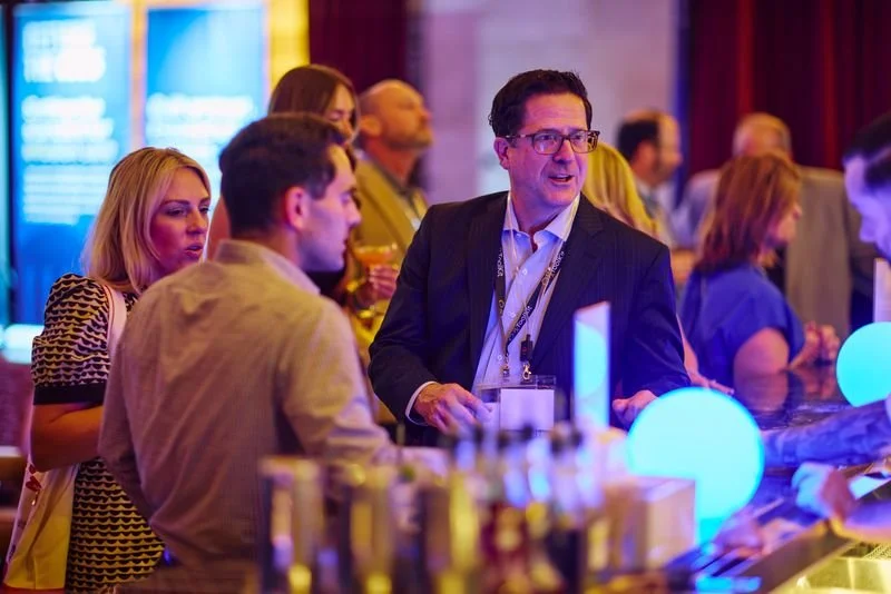 A man in a suit and glasses speaking to a group of people at a bar or social event, with women and men in the background, illuminated by blue and purple lighting.