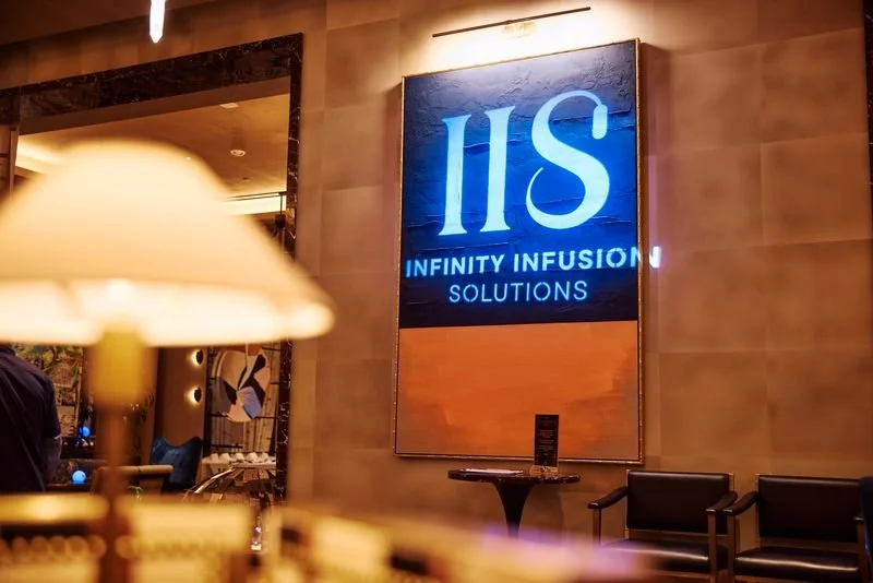 Interior of a lobby or reception area with a large illuminated sign reading 'ISIS INFINITY INFUSION SOLUTIONS' on the wall, with seating and a table nearby.