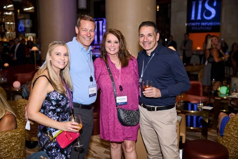 Four people standing together at a social event, smiling for the camera. Two women and two men, with drinks in hand, in a well-lit, lively indoor setting.