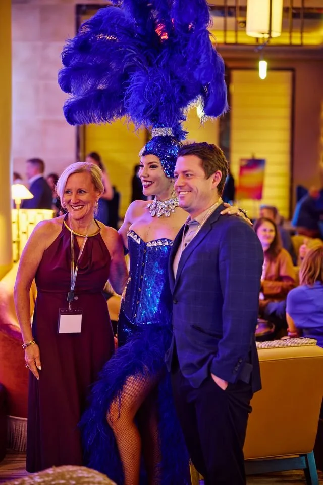 A performer in a blue sequined costume and large feathered headpiece standing between a smiling older woman in a maroon dress and a man in a dark suit, at a lively indoor event with warm lighting and people in the background.