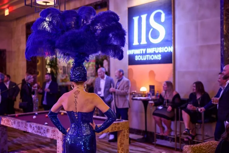 A woman wearing a blue sequined dress and large feathered hat dancing at an event, with a large sign for 'Infinity Infusion Solutions' on the wall and people socializing in the background.