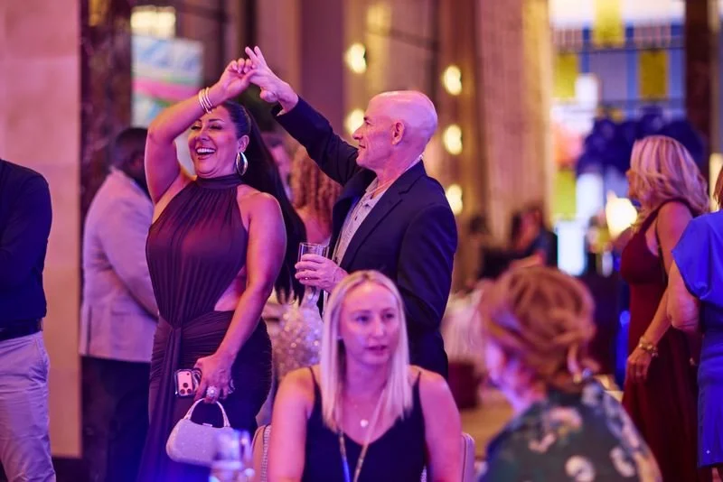 A woman in a purple dress dancing and smiling with a man in a suit at a party or celebration, surrounded by other guests.