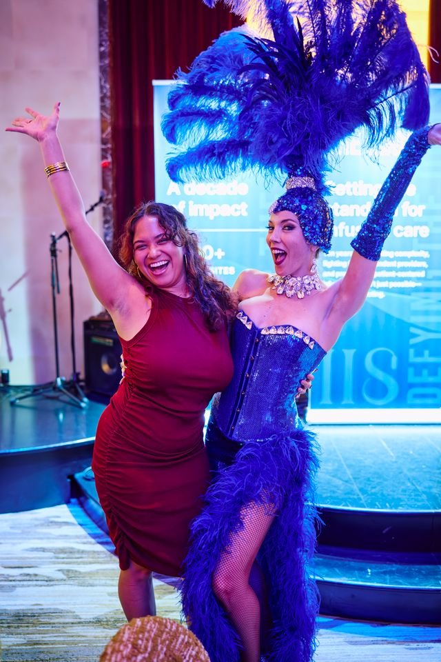 Two women: one in a red dress and the other in a blue showgirl costume with feathers, posing and smiling at an event.