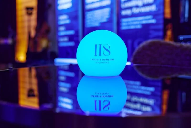 A glowing blue sphere with the text "IIS" and "Infinity Infusion Solutions" on a reflective surface at a conference or event.