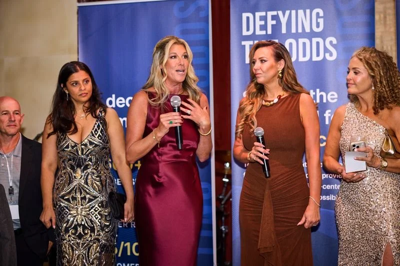 Four women standing in front of a blue backdrop at an event, with one woman speaking into a microphone, and others holding microphones or notes.