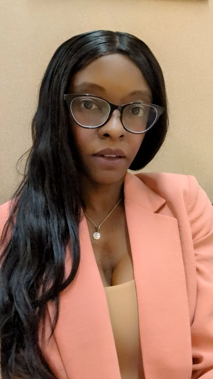 A woman with long dark hair, wearing glasses, a peach blazer, and a necklace, taking a selfie in front of a beige wall.