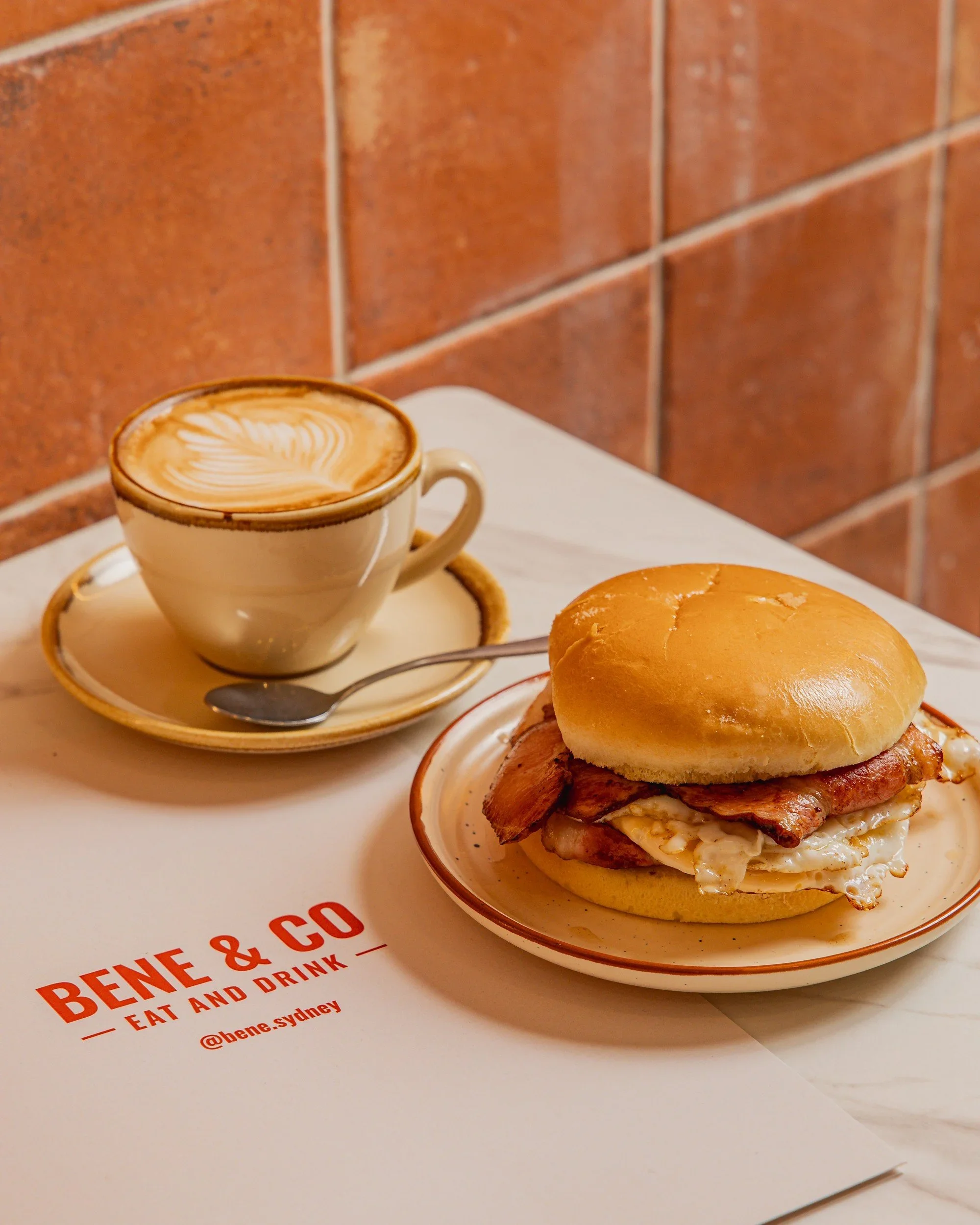 A bacon and egg roll on a ceramic plate next to a latte with latte art in a cream cup, placed on a marble table with the Bene & Co Eat and Drink logo, at a café in McMahons Point, North Sydney.