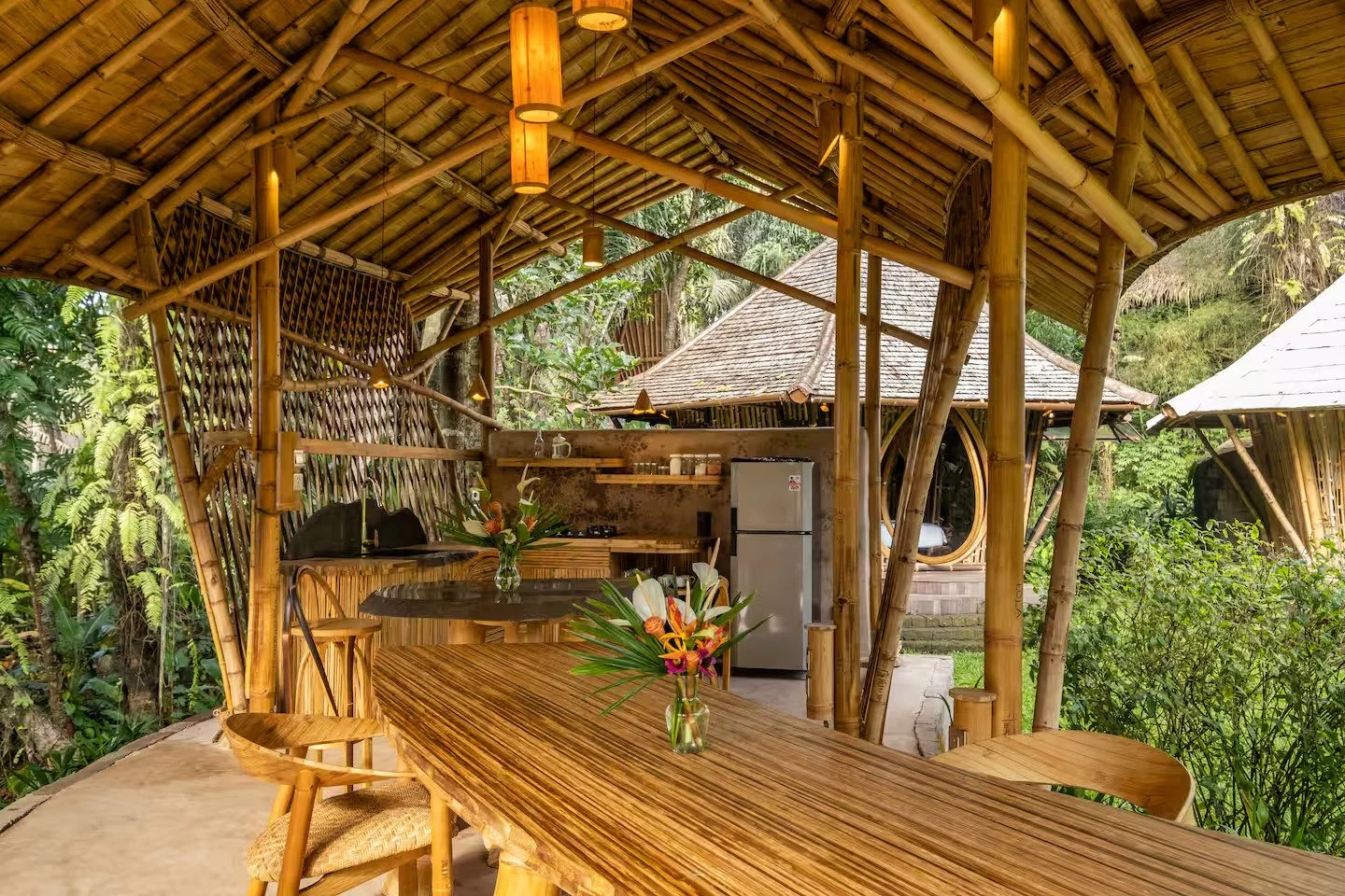 Open-air bamboo hut with a wooden dining table and chairs, decorated with flower vases, surrounded by greenery and other bamboo structures.