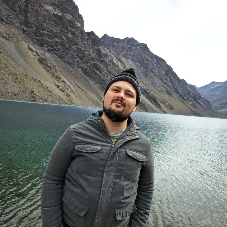 Homem de camiseta cinza e gorro preto em frente a um lago cercado por montanhas rochosas.