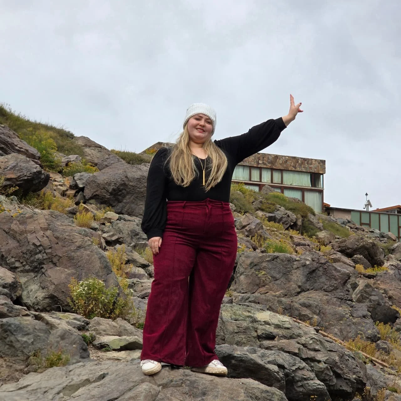 Mulher sorridente com cabelo loiro usando chapéu de lã branco, blusa preta e calças largas vermelhas, posando em uma área rochosa ao ar livre, com uma construção moderna ao fundo e céu nublado.