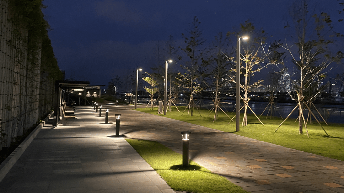 A nighttime view of a paved pathway along a river, illuminated by modern ground and street lights, with young trees supported by stakes lining the path, and a city skyline in the distance.