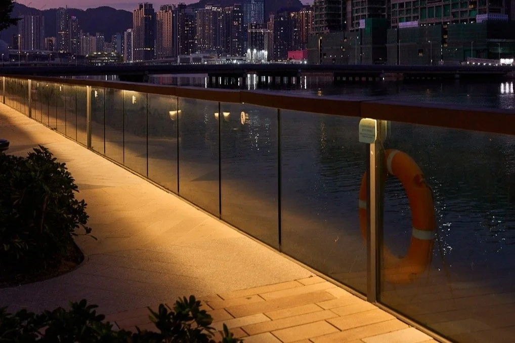Riverwalk promenade at night with glass railing, water reflecting city skyline, and a life buoy hanging on the railing.