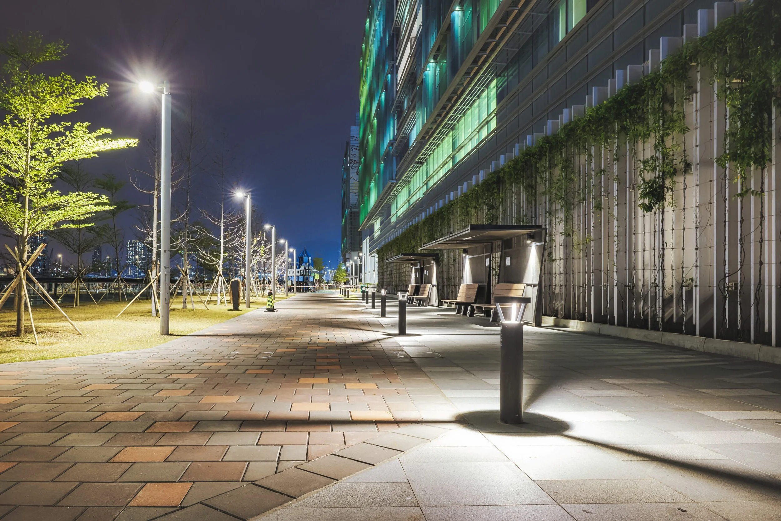 Nighttime view of a modern outdoor walkway with illuminated street lamps, benches, and trees, alongside a building with colorful lighting. City skyline visible in the distance.