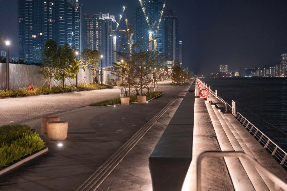 Night view of a waterfront promenade with streetlights, potted plants, trees, and a city skyline under construction with cranes in the background.