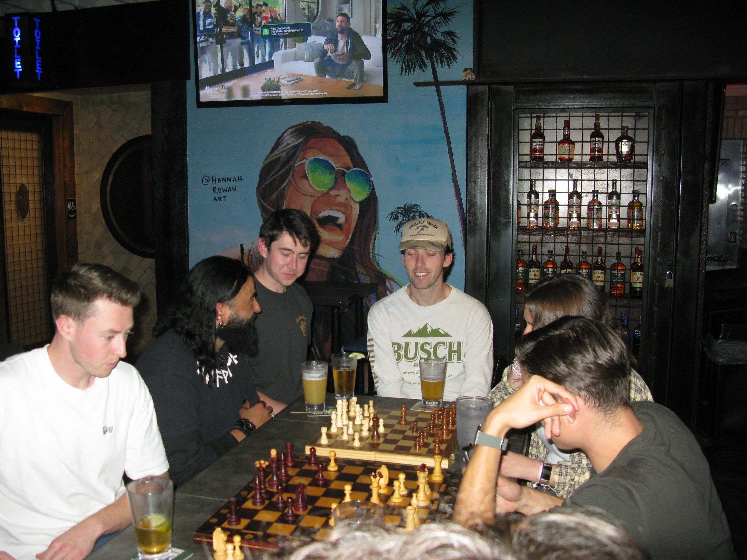 Group of people playing chess at a bar, with drinks on the table, and a colorful mural on the wall behind them.