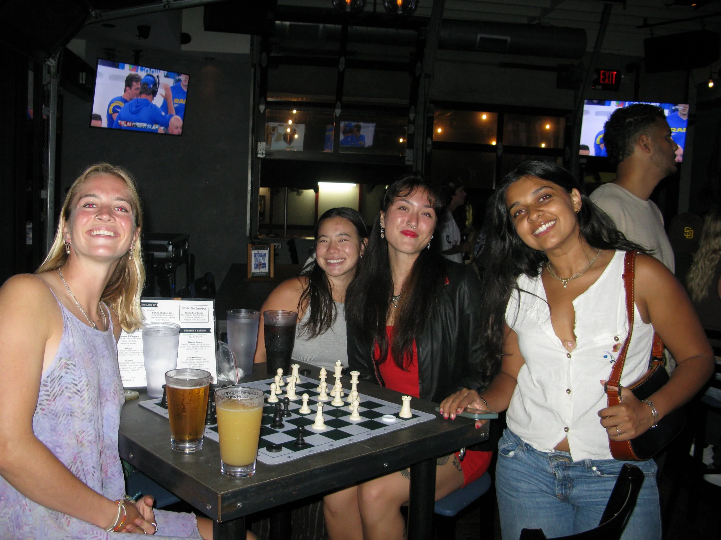 Four women sitting around a table with a chessboard, drinks, and a menu, smiling at the camera in a bar or pub.