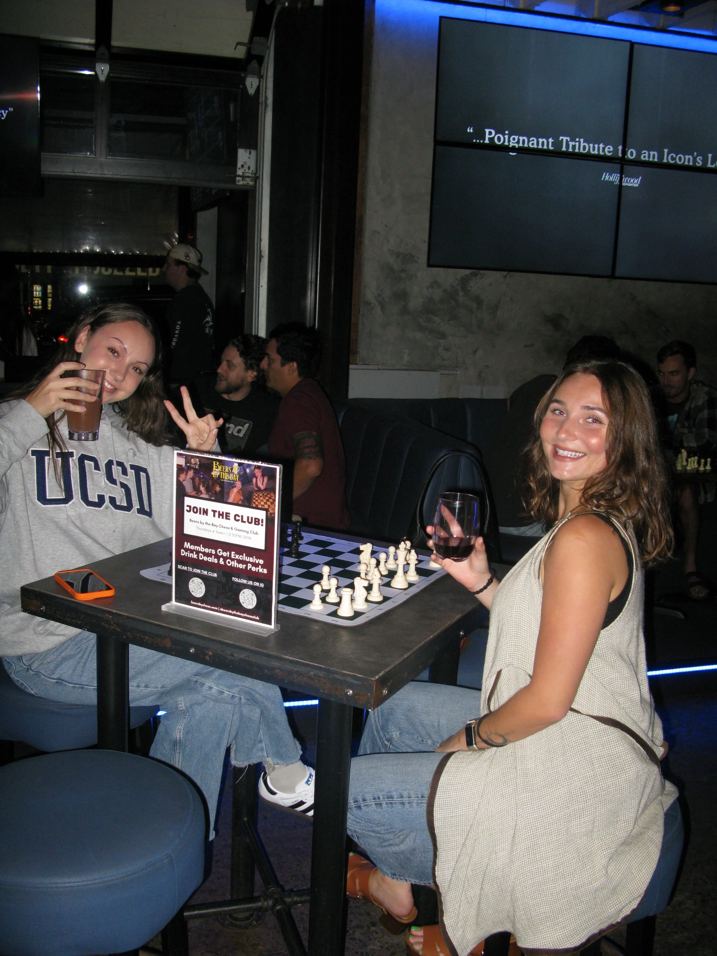 Two young women are sitting at a table playing chess at a bar or pub. One woman is wearing a grey UCSO sweatshirt, holding a drink in her hand, and making a peace sign. The other woman is wearing a beige sleeveless dress, holding a glass of red wine, and smiling. There are other people in the background, and a digital billboard or screen is visible on the wall.