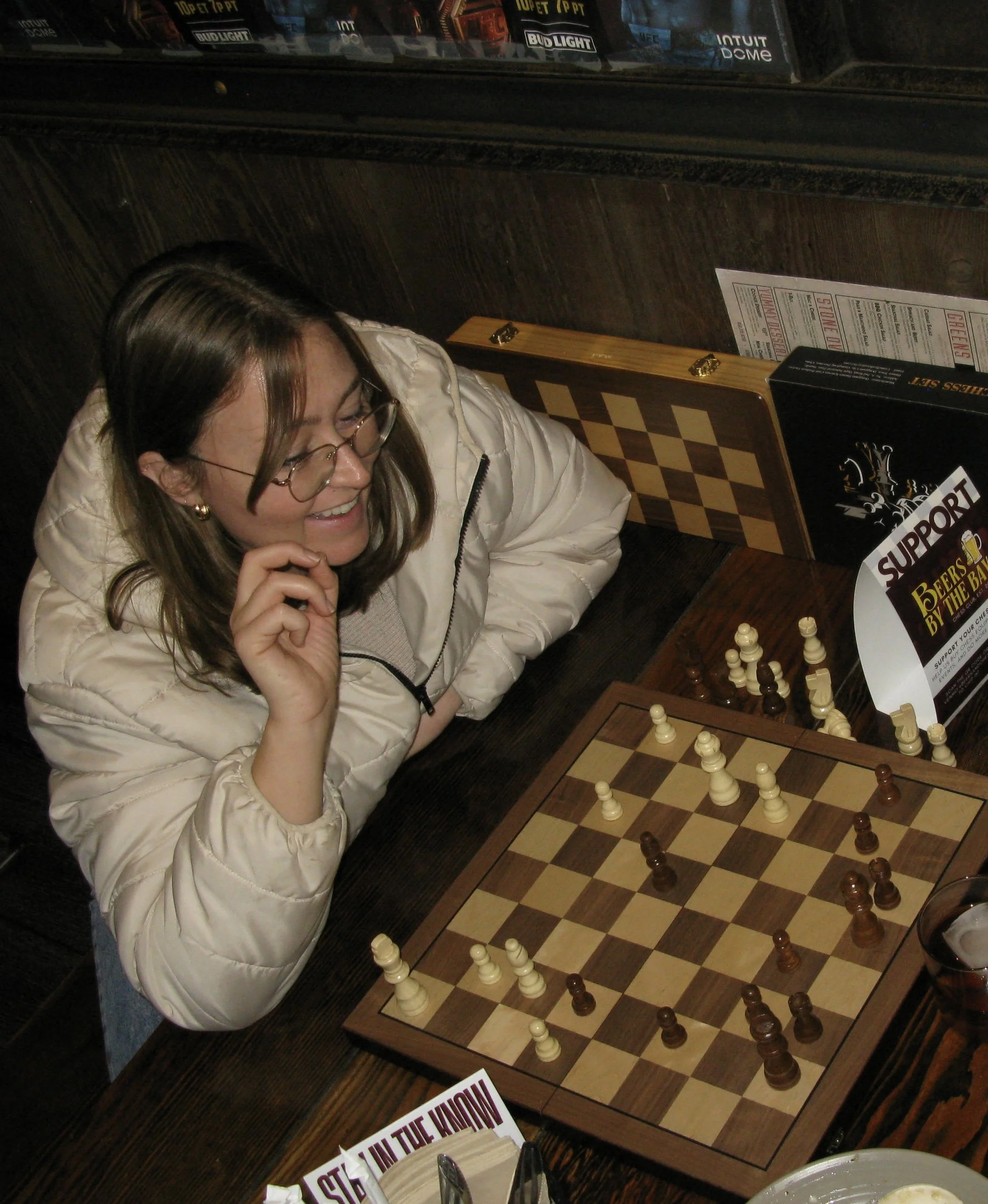 A woman with glasses and a beige jacket playing chess at a table in a restaurant or bar.