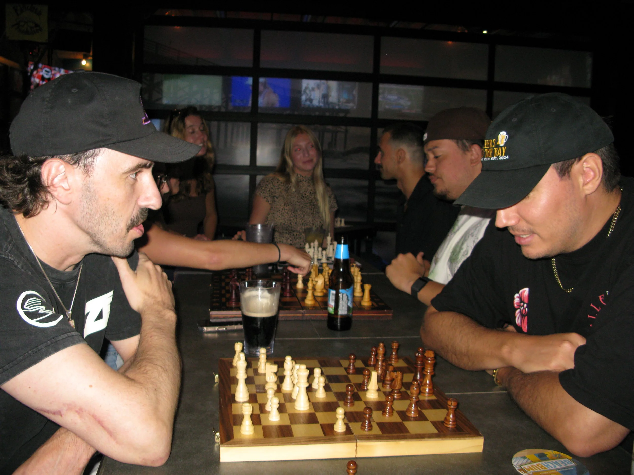 Two men playing chess at a bar or restaurant, with multiple other people watching and socializing in the background.